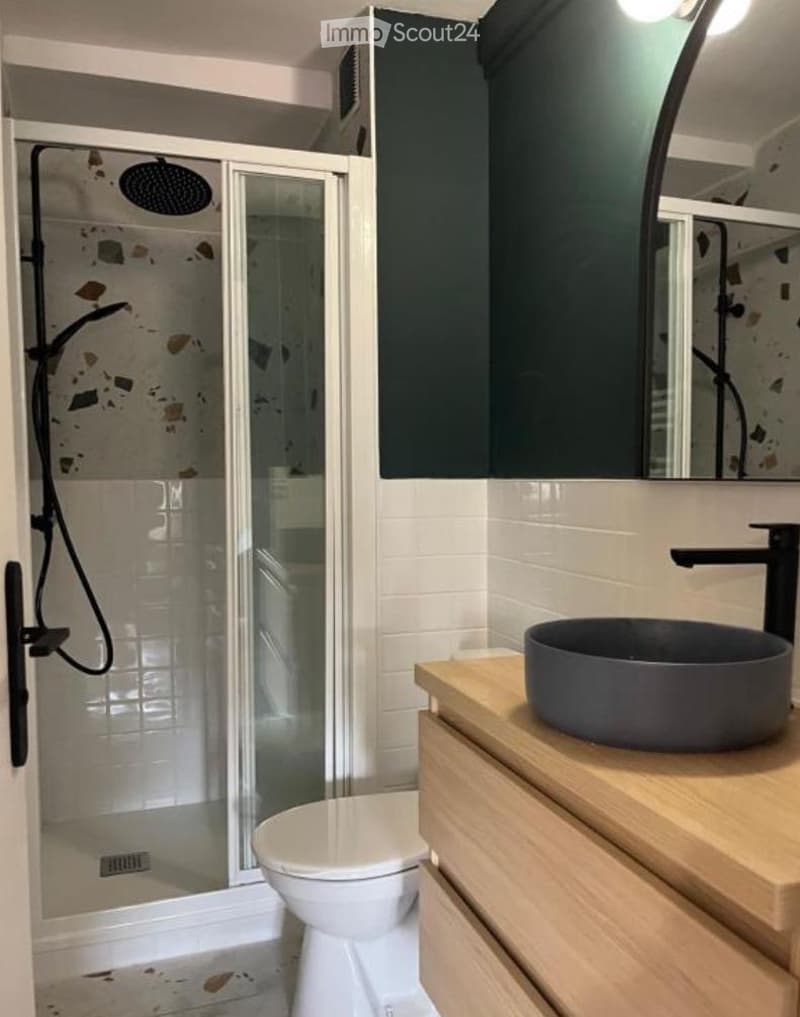 Modern bathroom with a white toilet, dark grey sink, wooden vanity, large mirror, and a glass-enclosed shower with a modern showerhead.