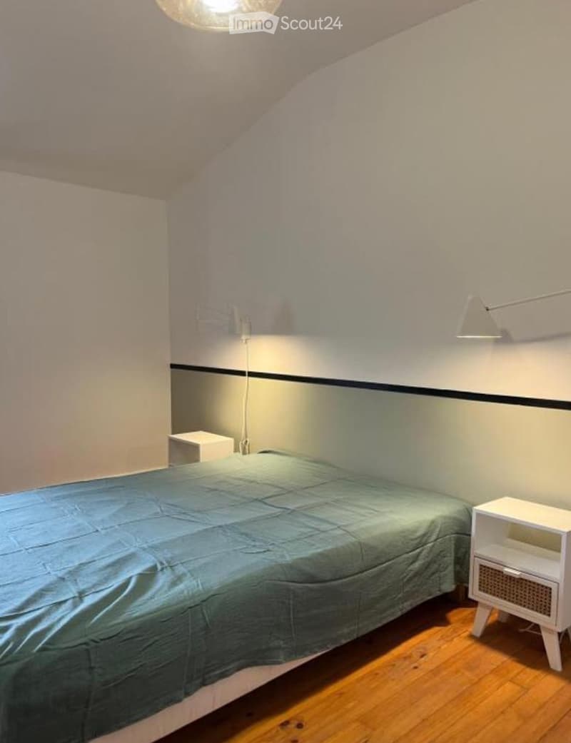 Cozy bedroom with a single bed, nightstand, and wall-mounted lamp. The room features wooden flooring and a slanted ceiling.