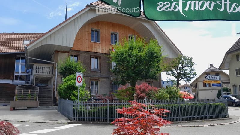 Two-story residential building with a wooden facade, tiled roof, and a small garden area enclosed by a metal fence.
