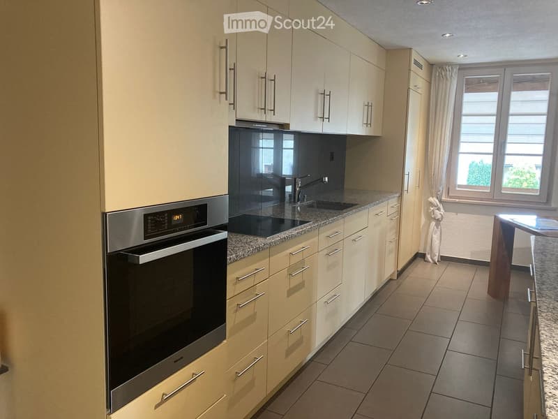 Modern kitchen with light-colored cabinets, granite countertop, built-in oven, sink, and faucet. Features a window with curtains and a small table near the window.