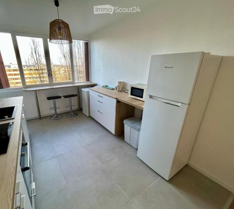 Modern kitchen with white cabinets, a refrigerator, microwave, and a kitchen island with stools. Large windows provide natural light.