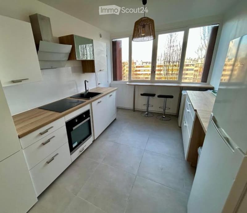 Modern kitchen with white cabinets, wooden countertops, built-in oven, sink, and stools at a small dining table. Large windows provide natural light.