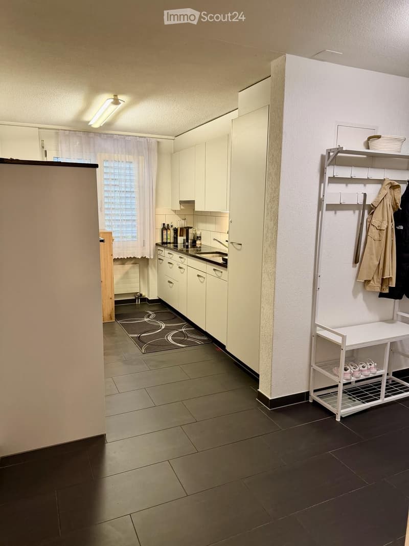 A modern, well-lit kitchen with white cabinets, a sink, and a small shelf holding coats and shoes. The floor is tiled, and there is a window with a curtain.