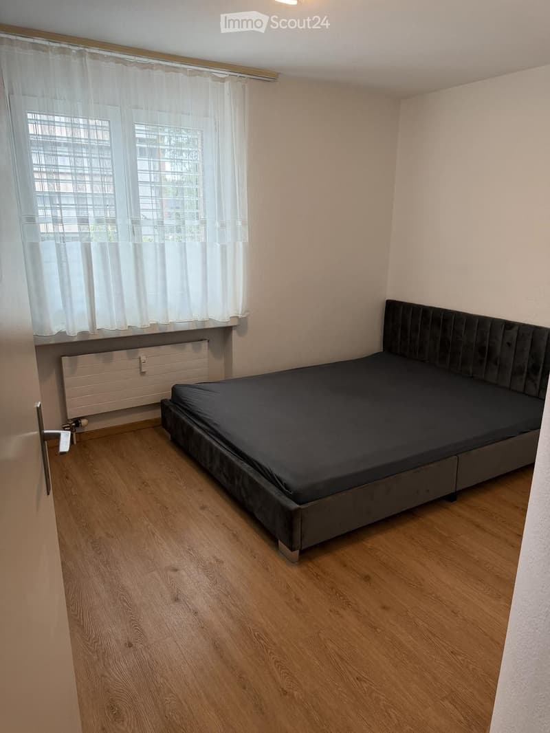 A small bedroom with a single bed, wooden flooring, and a window with white curtains. The room has a radiator below the window and a door on the left side.