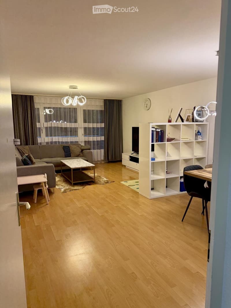 Modern living room with wooden flooring, large windows, a grey sofa, a coffee table, a shelf with books and decorative items, a dining table with chairs, and a small stool near the sofa.