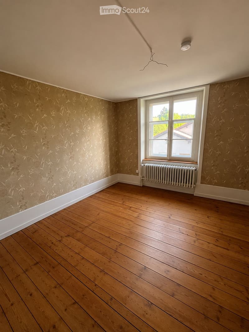 Empty room with wooden flooring, patterned wallpaper, a single window with white frame, and a radiator below it.
