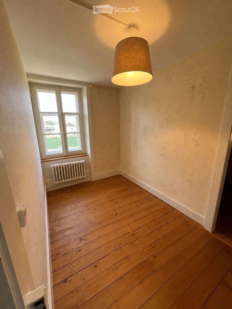 Empty room with wooden flooring, white walls, a window with a radiator below, and a hanging light fixture.