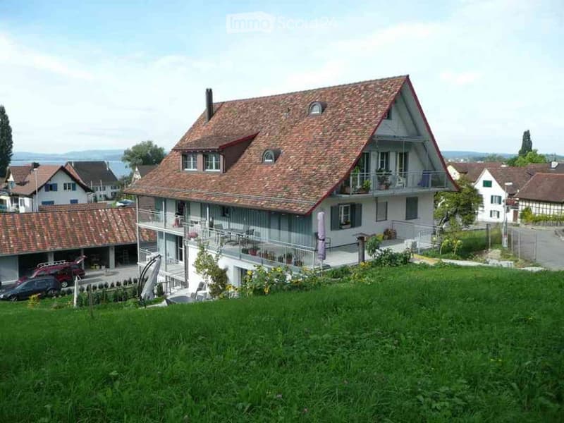 Two-story house with a red-tiled roof, white walls, and a balcony overlooking a green lawn. Garage and parking area visible in the foreground.