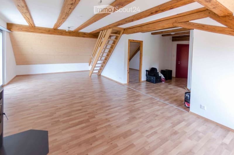 Empty room with wooden flooring and exposed wooden beams on the ceiling. A wooden staircase leads to an upper level, and there are a few storage items in the corner.