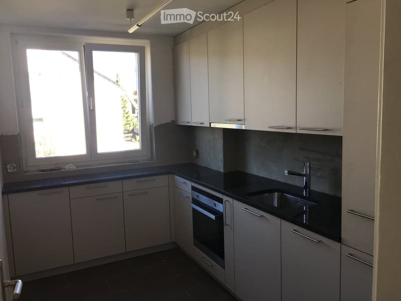 Modern kitchen with white cabinets, dark countertops, and stainless steel appliances including a sink, faucet, and oven. Large windows provide natural light.