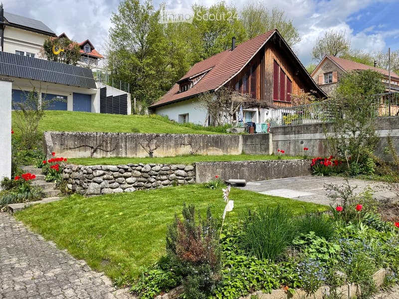 Well-maintained garden with green lawn, stone pathways, flower beds, and surrounding houses with sloped roofs and solar panels.