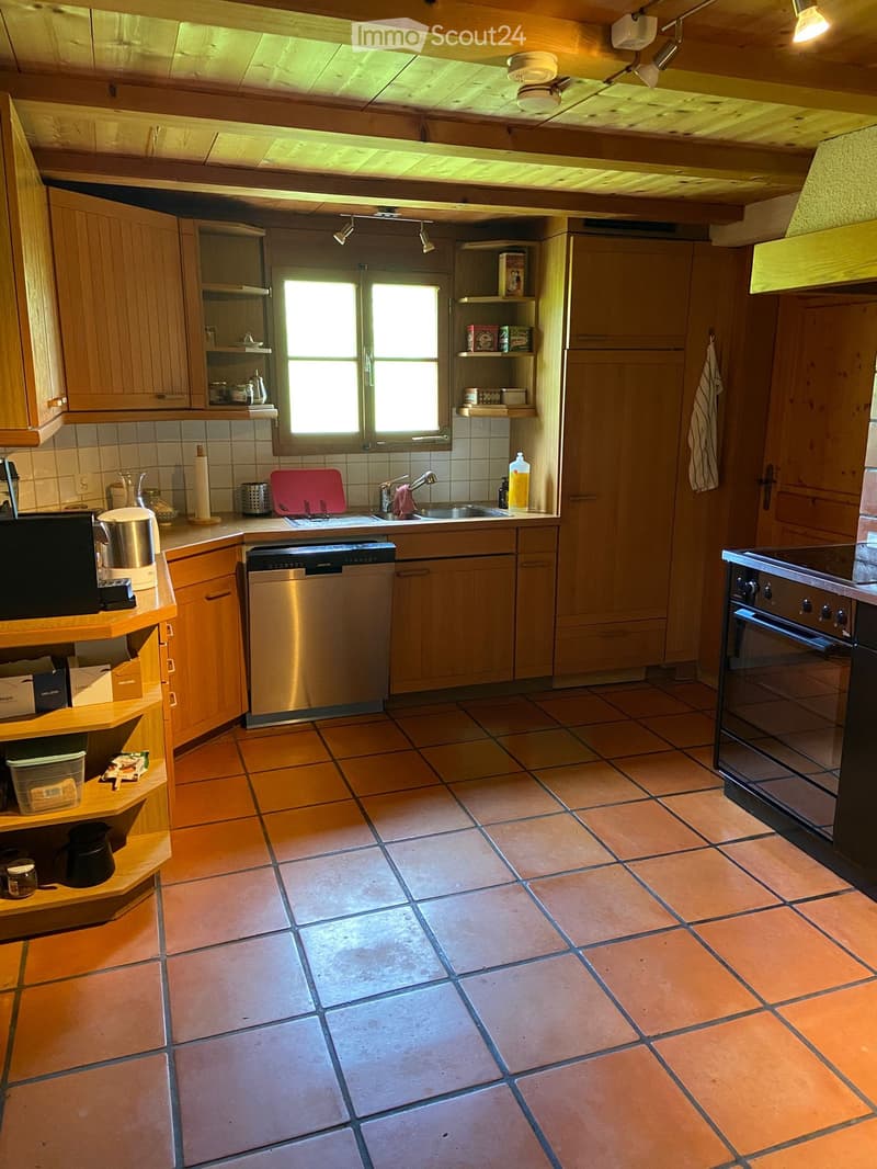 The kitchen features wooden cabinets, a tiled floor, and stainless steel appliances including a sink, oven, and dishwasher. There is a window above the sink, and various kitchen items are placed on the countertops and shelves.