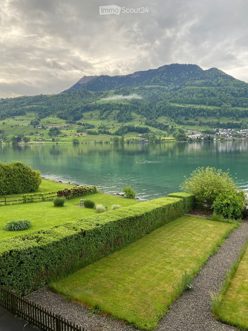 A serene garden area with neatly trimmed hedges, a gravel pathway, and a view of a calm lake surrounded by lush green hills and a distant mountain.