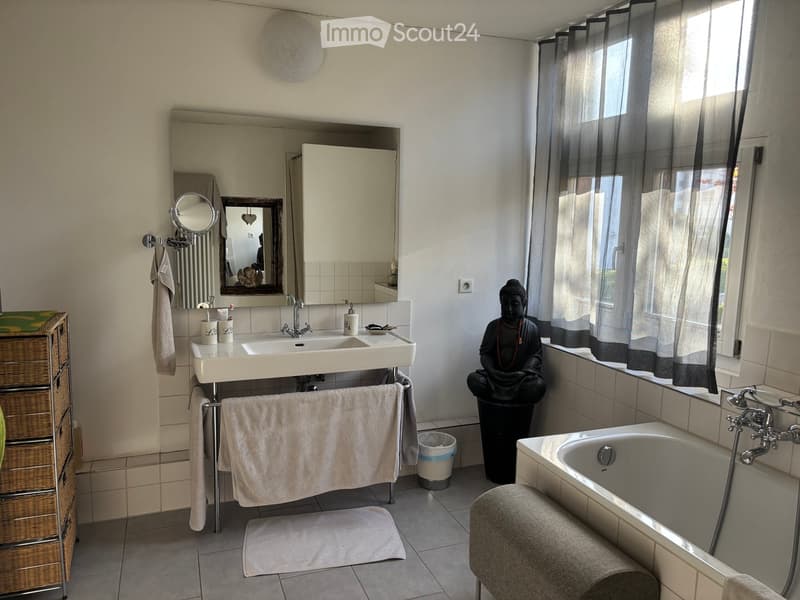 white bathtub, vanity with double sinks, large mirror, toilet, Buddha statue, small cabinet, shelves, curtained window, towel rack