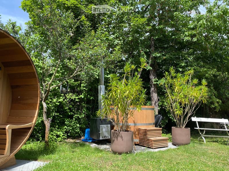 A garden with a wooden sauna, a barrel, a bench, and potted plants on the grassy lawn.