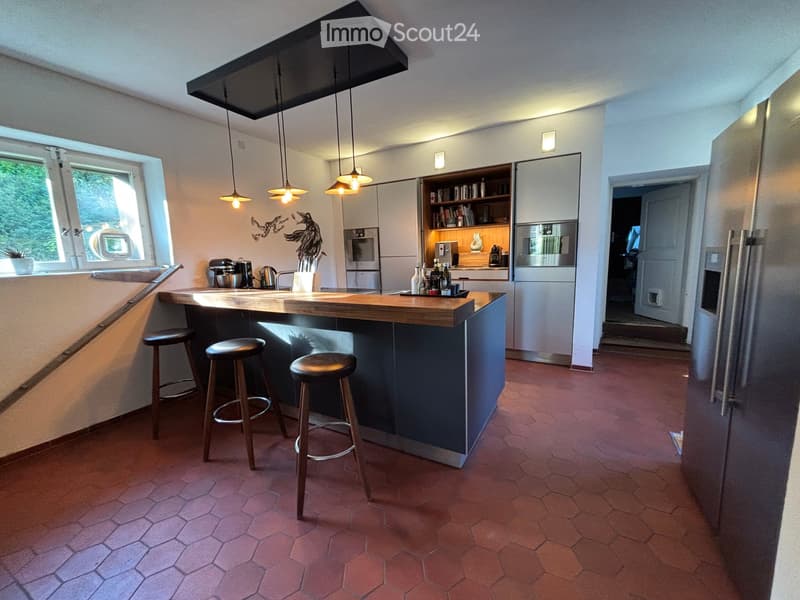 Open kitchen with wooden countertops, stainless steel backsplash, tiled floor, hanging lamps, kitchen island with stools, stainless steel appliances, built-in cabinets, microwave, coffee machine, dining area with table and chairs, window with outdoor view