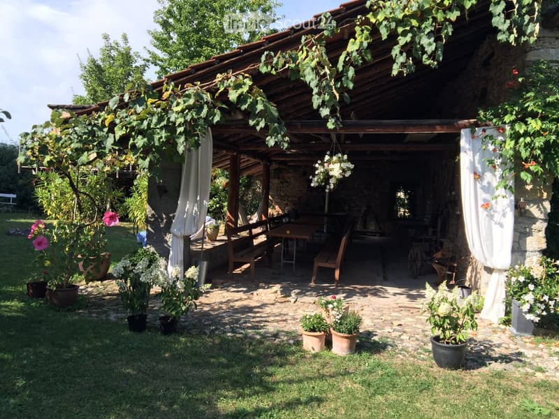 Rustic building with stone walls and wooden pergola, outdoor dining area, wooden benches, table, hanging plant pots, vine covering the pergola, fireplace.