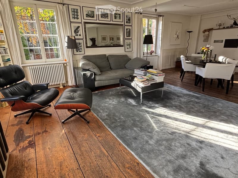 Modern living room with a view to the street, wooden floor, large windows, gray couch, a black chair, coffee table, a stack of books, a gray carpet, dining table and chairs.
