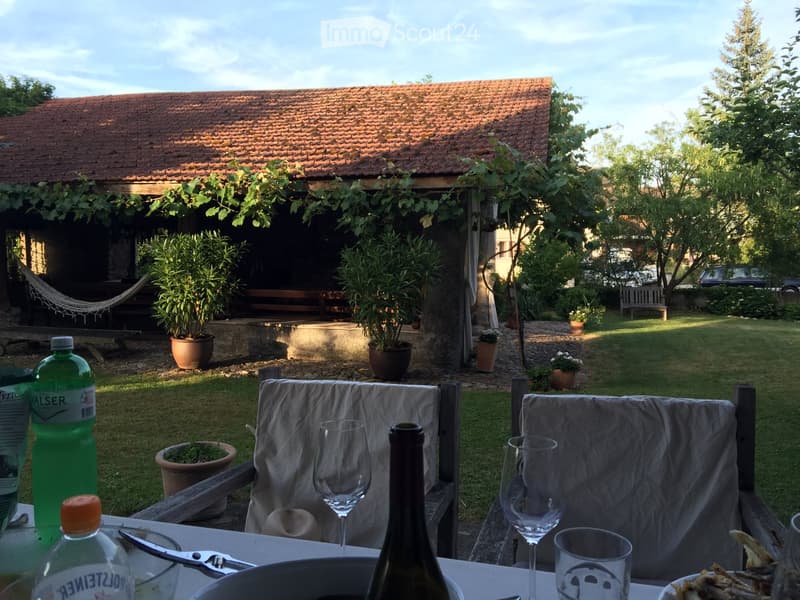 House with red roof, hammock, several potted plants, wooden benches, garden, table with bottles, glasses, wine glasses, food