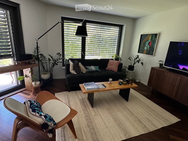 Modern living room with a black sofa, wooden coffee table, armchair, floor lamp, TV, and decorative plants.