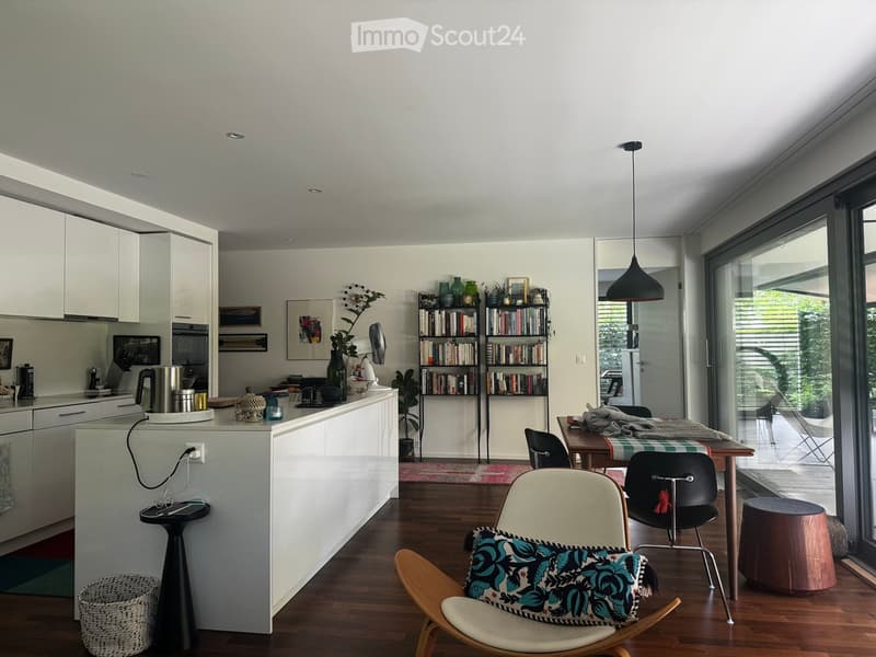 Modern open-plan living and dining area with a kitchen island, white cabinets, pendant light, bookshelf, and various seating options including a sofa, chairs, and a stool.