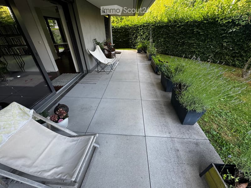 Modern balcony with concrete floor, lounge chairs, small table, and potted plants.