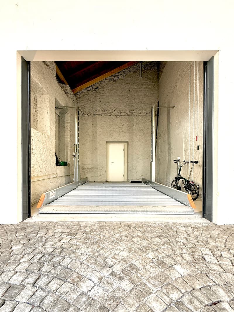 An outdoor area with a raised ground floor leading to a covered entrance. The entrance features stone walls, a small white door, and a bicycle parked inside. The ground is paved with cobblestones.