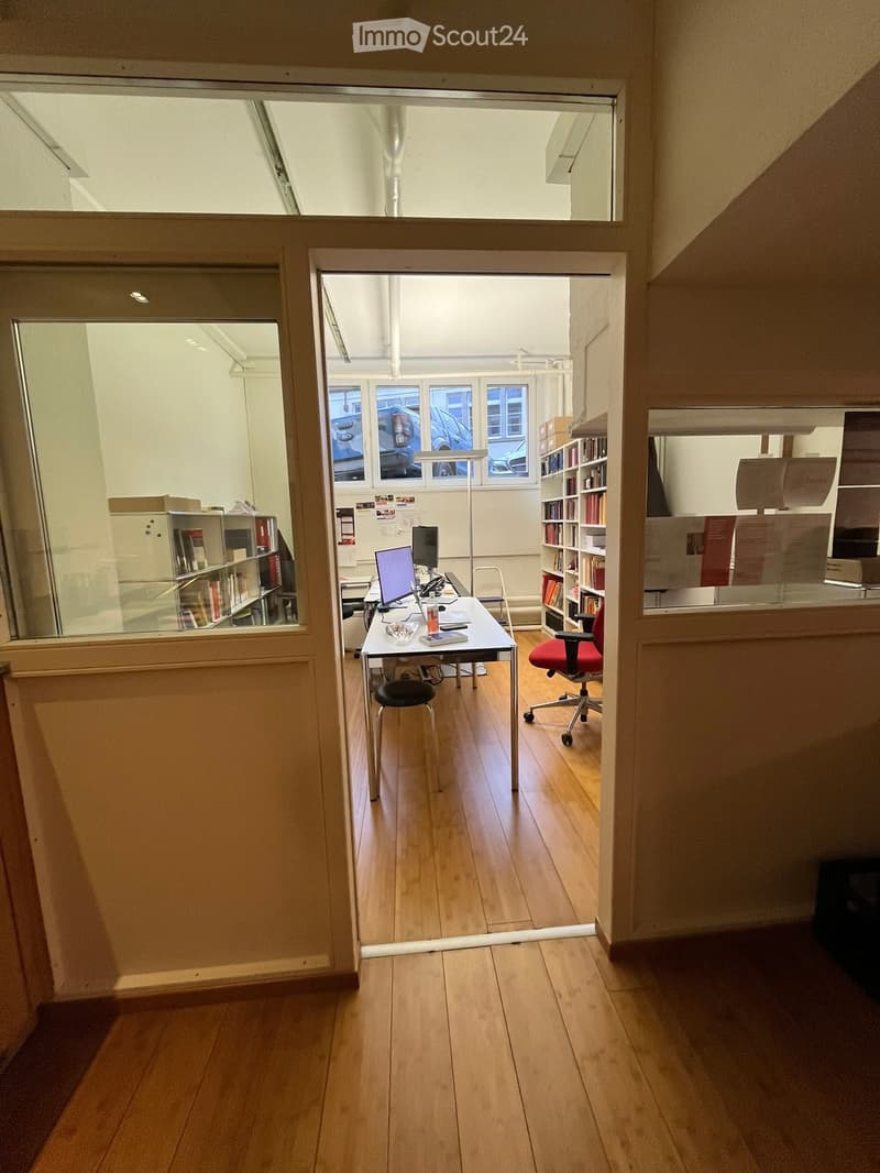 Open door with wooden floor, desk with monitor, red chair, book shelves, office supplies