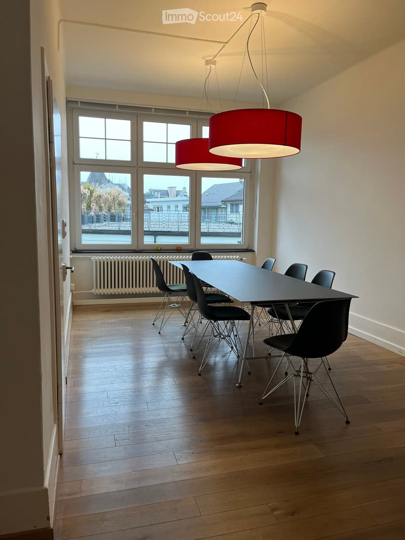 Meeting room with white walls, wooden floor, a large table, black chairs, two hanging lamps, a radiator, and a large window.