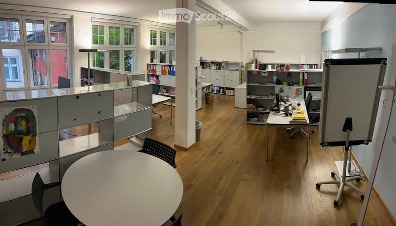 An empty office space with round table, several black chairs, cabinets, bookcases, and multiple computer equipment. There are windows with trees outside, and the room has a wooden floor.