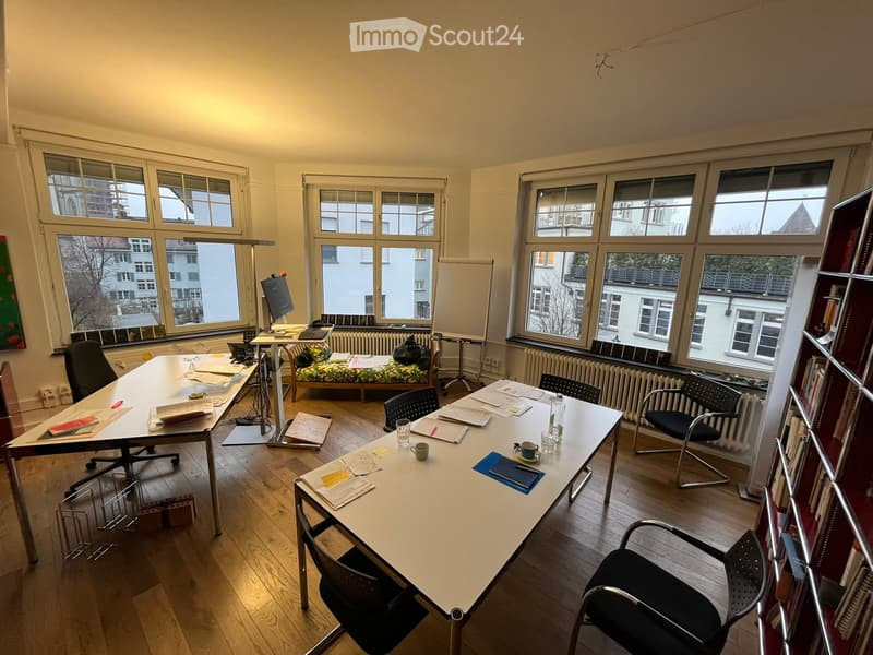 an office space with large windows, a radiator, two desks, six chairs, a couch, a bookshelf, a monitor and keyboard