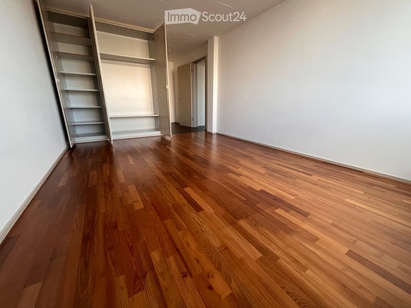 Empty room with wooden flooring and white walls, featuring an open closet with empty shelves and a doorway leading to another room.