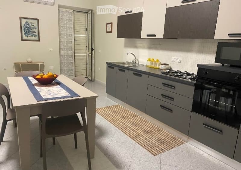 Modern kitchen with grey cabinets, stainless steel appliances, a dining table with chairs, and a bowl of fruit on the table.