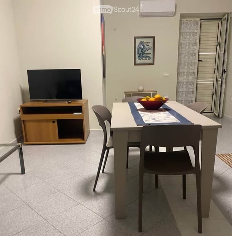 A modern dining area with a white table, four chairs, a TV on a wooden stand, and a bowl of fruit on the table. The room has tiled flooring and an air conditioning unit on the wall.