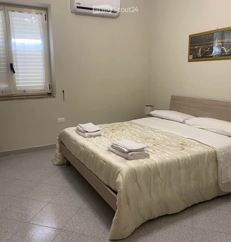 A neatly made double bed with white pillows and folded towels on top, placed against a light-colored wall with a window and an air conditioning unit above it. The room has tiled flooring and a framed picture on the wall.