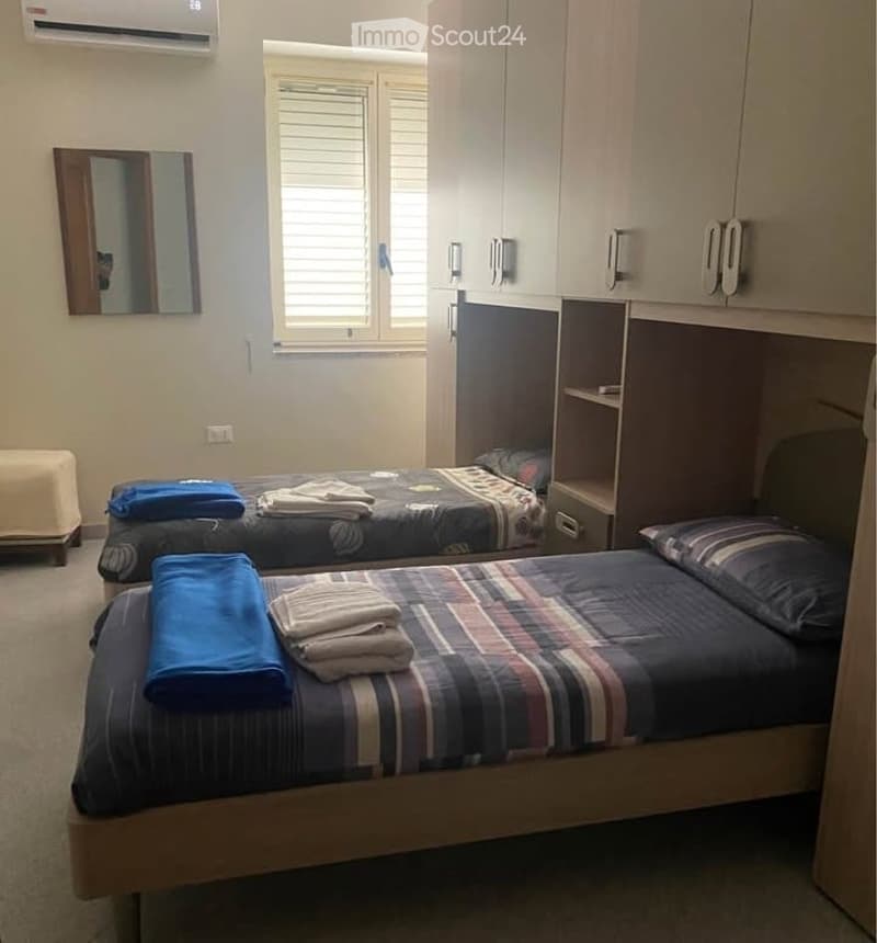 Cozy bedroom with two single beds, neatly made with patterned bedspreads and folded towels, a mirror on the wall, built-in cabinets, and a small stool near the window.