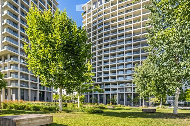 Modern high-rise residential building with multiple balconies and large windows, surrounded by green spaces and trees.