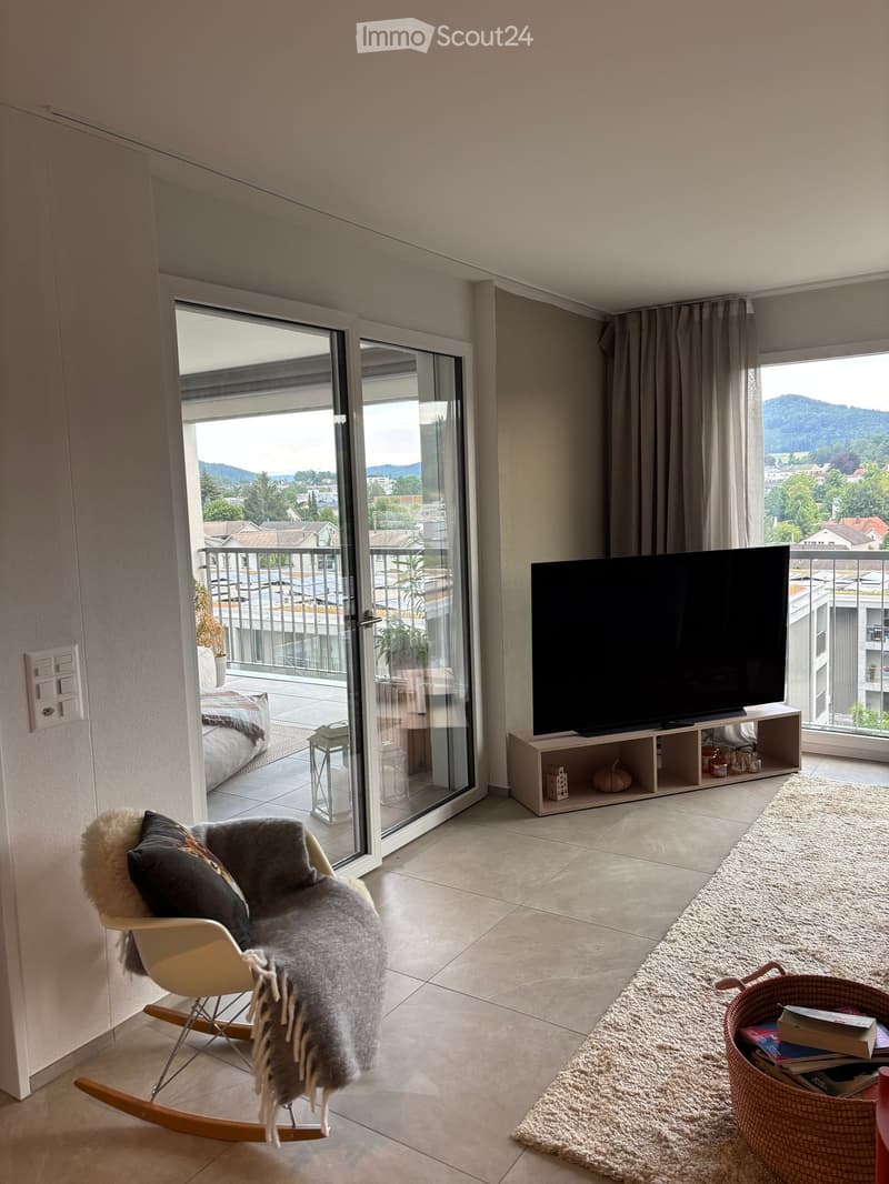 Modern living room with large windows and a view, featuring a sofa, television, and a small table.