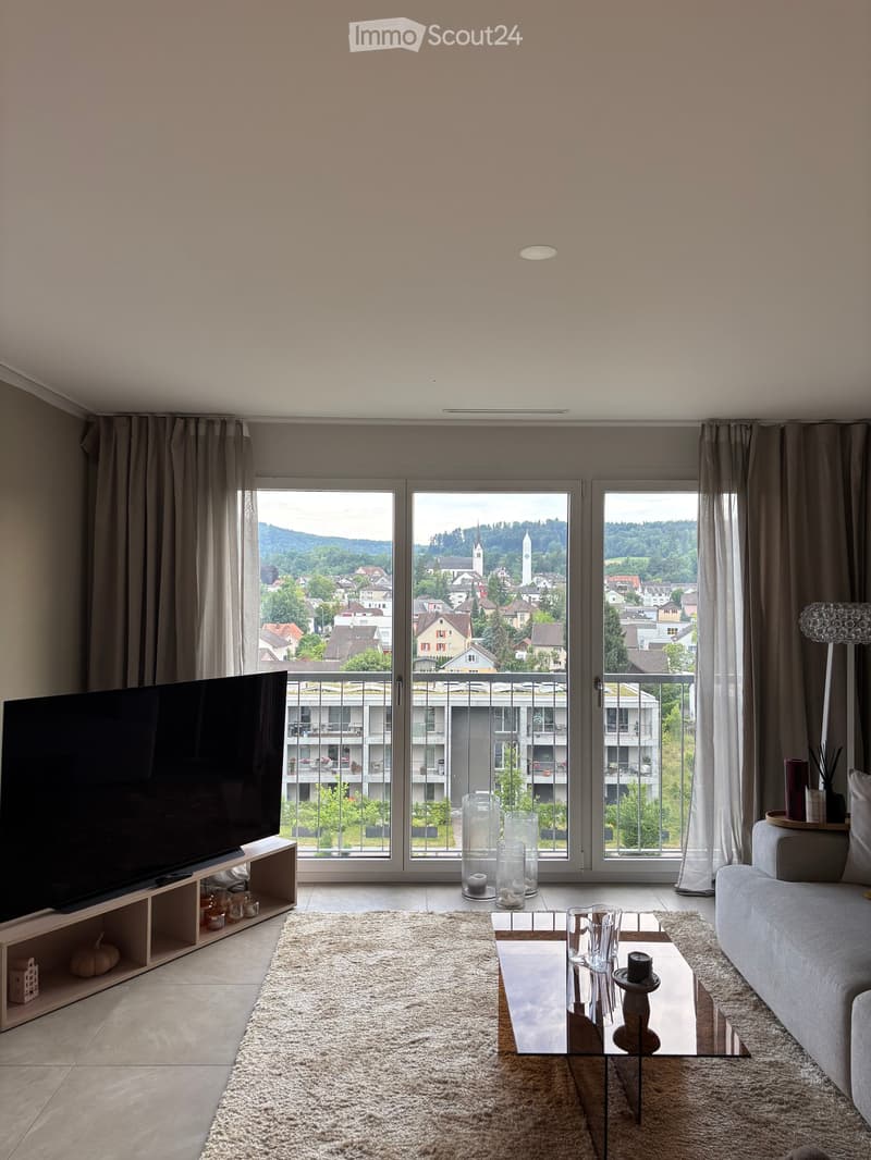 Modern living room with large windows offering a scenic view, featuring a sofa, coffee table, television, floor lamp, and decorative vases.