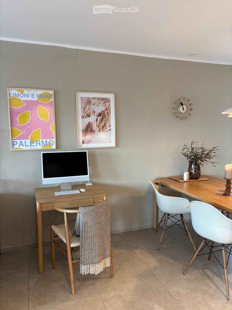 A modern, well-lit room with two desks, one with a computer setup and the other with a vase and candle, featuring contemporary chairs and artwork on the walls.