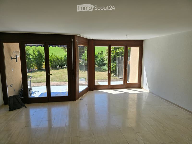 Empty room with tiled floor, large sliding glass doors leading to a garden, and a wall-mounted light fixture.