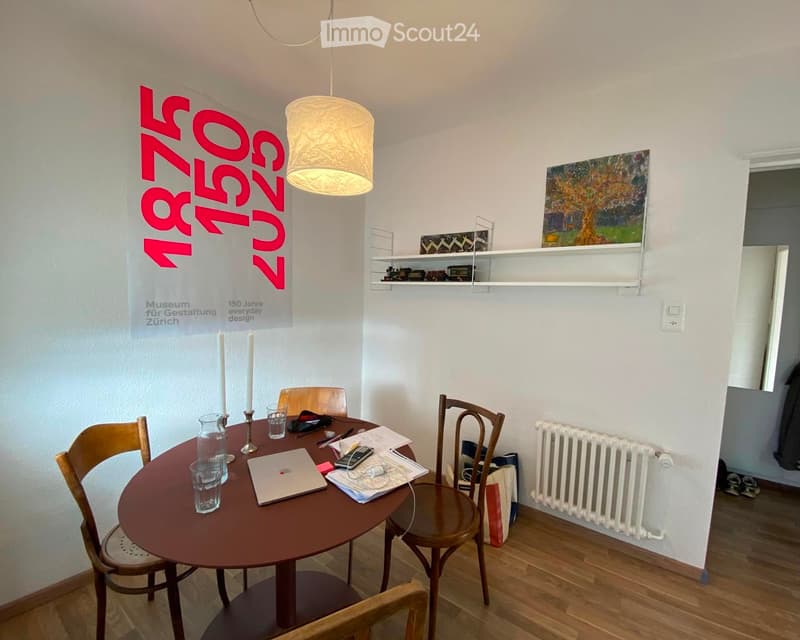 A small, well-lit room with a round wooden table surrounded by four wooden chairs. The table holds a laptop, a vase, and some papers. A white radiator is mounted on the wall, and a painting hangs above a shelf with small decorative items. A banner with te