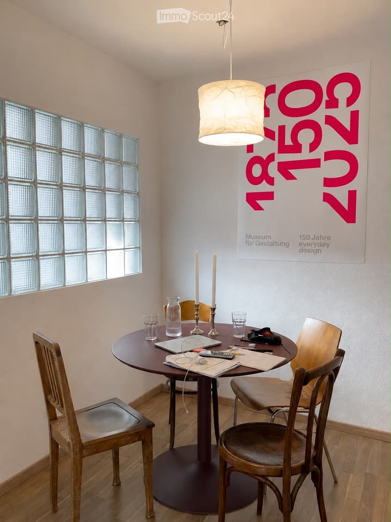 A modern dining area with a round wooden table, four wooden chairs, a laptop, candles, a vase, and cups.