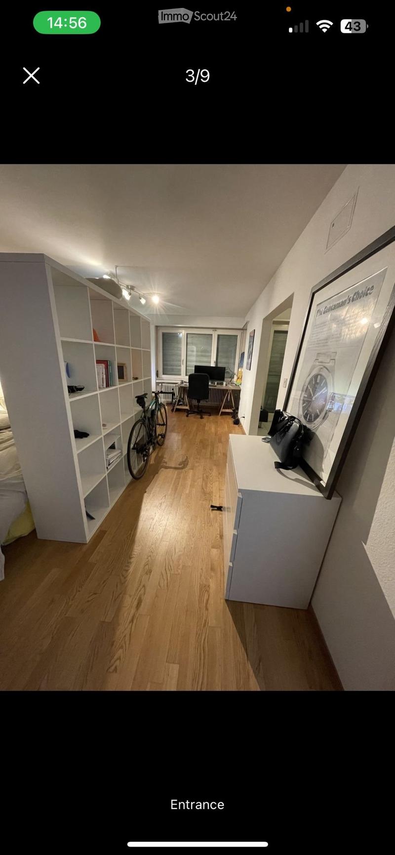 A furnished room with wooden flooring, a bicycle, a desk with a laptop, a chair, a shelf, and a framed picture on the wall.
