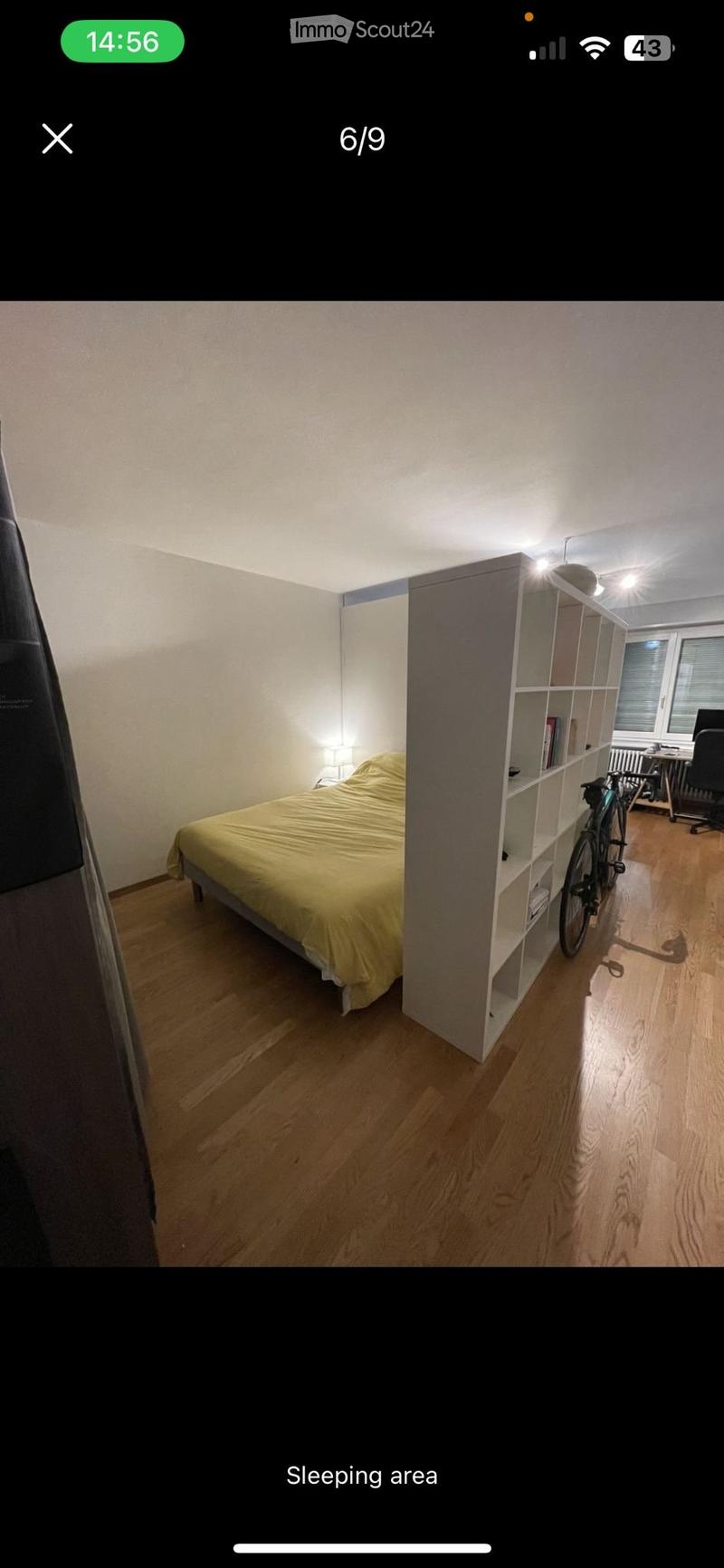 A modern bedroom with a wooden floor, a bed with yellow bedding, a nightstand with a lamp, a bookshelf, and a bicycle leaning against it.