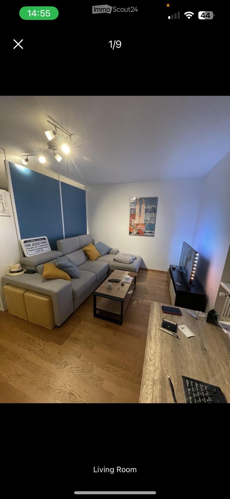 Modern living room with a grey sectional sofa, wooden coffee table, TV stand, and a small desk with a laptop and keyboard. The room features wooden flooring, a blue accent wall, and decorative artwork.