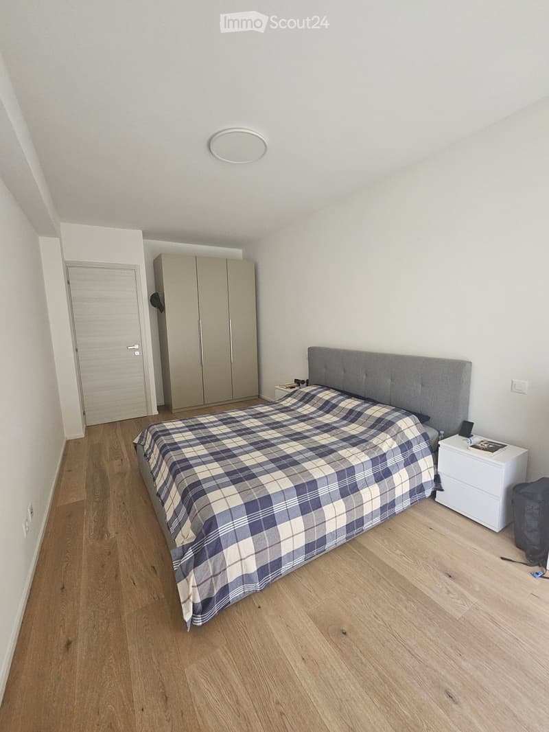 Modern bedroom with wooden flooring, a large bed with a grey headboard and plaid duvet, a white nightstand, and a wardrobe with mirrored doors.