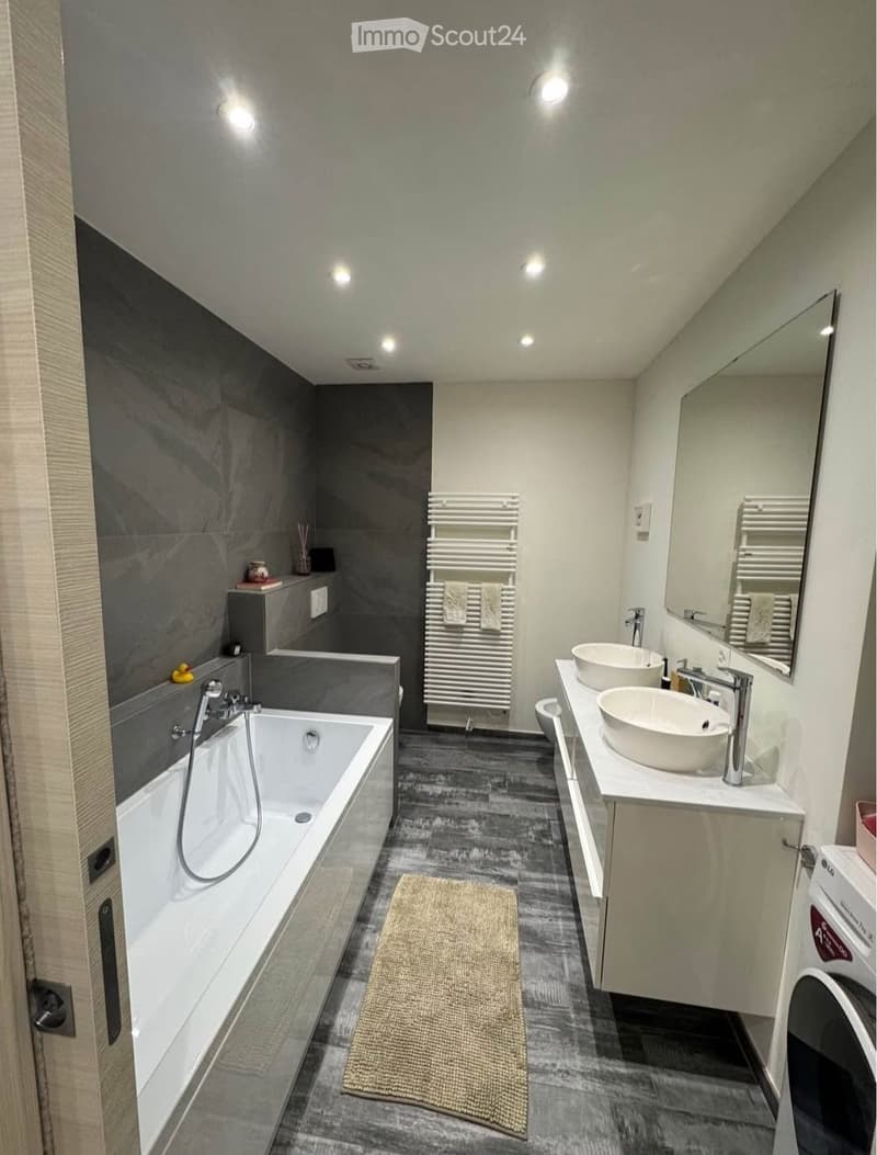 Modern bathroom with a bathtub, double sink, large mirror, radiator, and a small washing machine. The walls are tiled in dark grey, and the floor is dark grey with a beige rug.