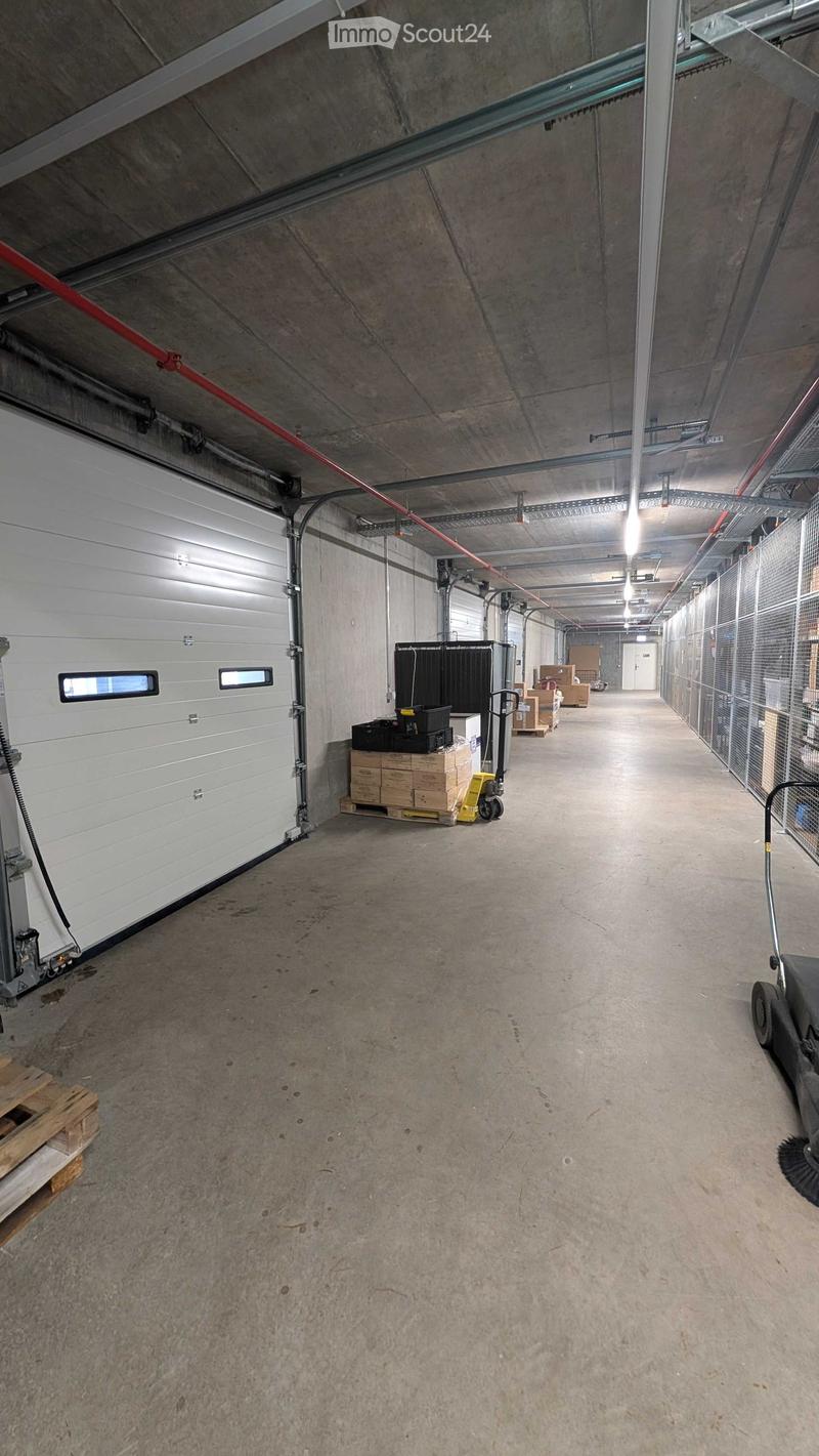 A long, wide concrete corridor with a high ceiling, industrial lighting, and a large garage door on the left. Storage items and a pallet are visible near the garage door.