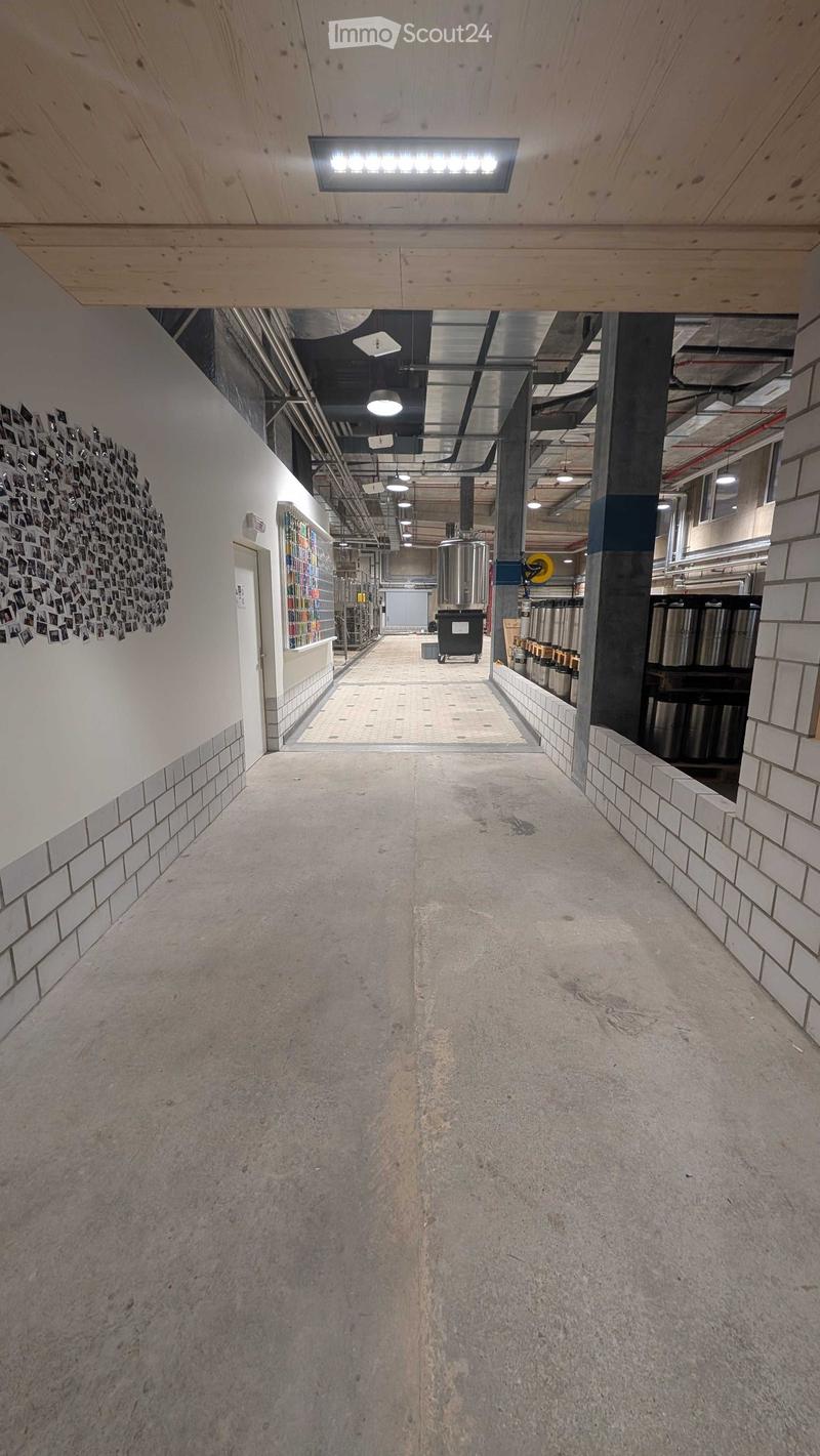 A long, industrial-style corridor with exposed pipes, concrete floors, and tiled walls. Features include a large circular photo collage on the left wall, a display shelf, and several large metal tanks in the background.